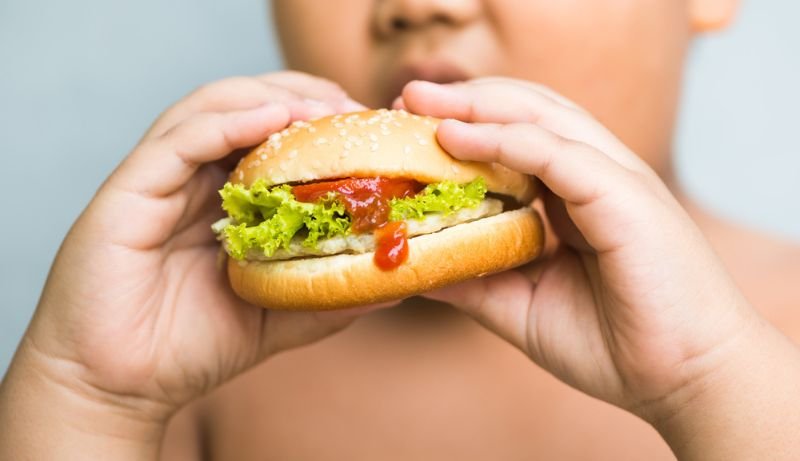 crianca com disturbio de obesidade infantil segurando hamburguer com as duas maos e o levando a boca